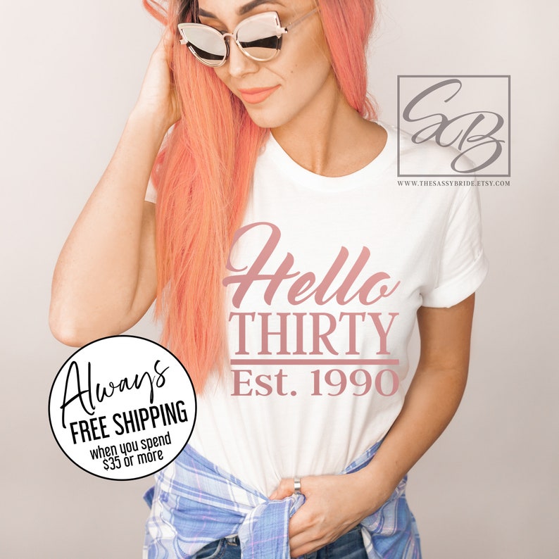 May include: White t-shirt with the text "Hello Thirty Est. 1990" in pink lettering. The shirt is being worn by a person with pink hair.