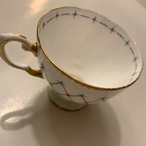Puede incluir: Taza de t&eacute; blanca con ribete dorado y asa decorativa. La taza presenta un patr&oacute;n geom&eacute;trico con detalles turquesa y rojos. El borde y la base tambi&eacute;n est&aacute;n adornados con dorado, creando un aspecto elegante.