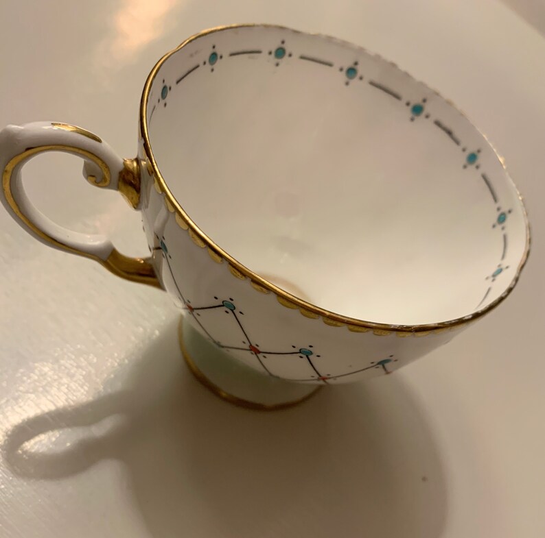 Puede incluir: Taza de t&eacute; blanca con ribete dorado y asa decorativa. La taza presenta un patr&oacute;n geom&eacute;trico con detalles turquesa y rojos. El borde y la base tambi&eacute;n est&aacute;n adornados con dorado, creando un aspecto elegante.