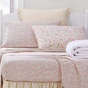 May include: A bed with floral print bedding, including a fitted sheet, flat sheet, and pillowcases. The sheets have a pink and green floral pattern on a white background. A folded white blanket and a gingham pillow are also visible.