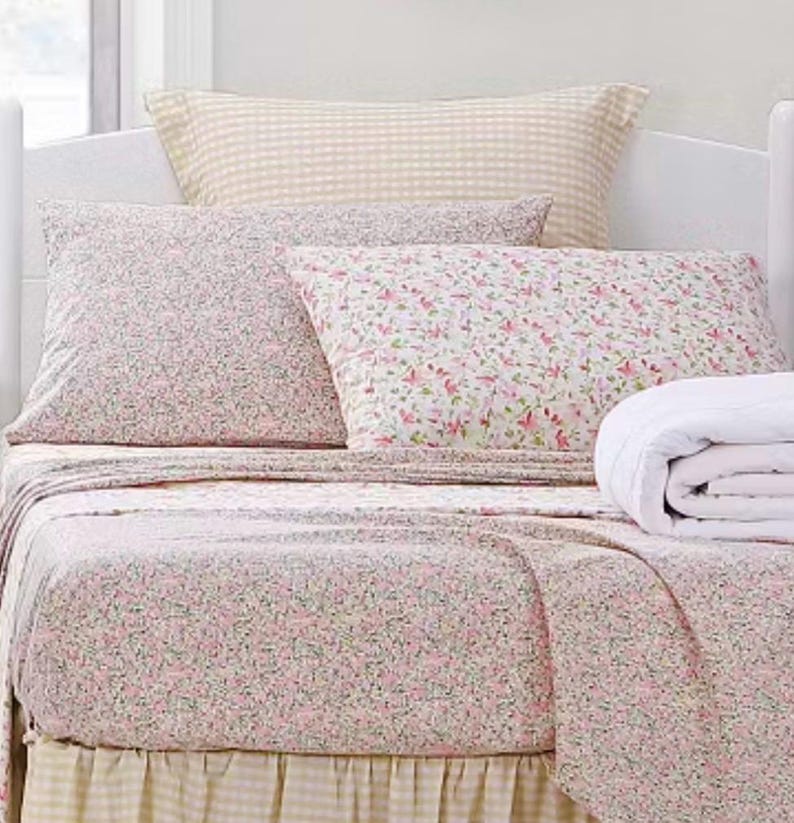 May include: A bed with floral print bedding, including a fitted sheet, flat sheet, and pillowcases. The sheets have a pink and green floral pattern on a white background. A folded white blanket and a gingham pillow are also visible.