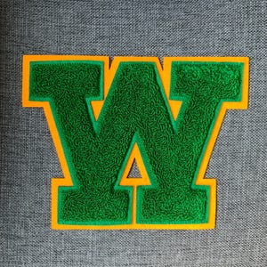 May include: A green chenille letter "W" patch with an orange border. The letter is on a gray fabric background. The patch is likely for a jacket or bag.