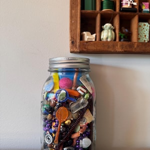 May include: A large glass jar filled with a variety of small, colorful objects, including beads, a miniature tennis racket, and other trinkets. The jar has a silver lid and sits on a gray surface. A wooden shelf with small items is in the background.