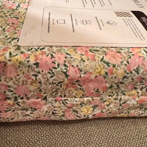 May include: A floral patterned fabric, featuring pink, yellow, and green flowers on a white background. The fabric is folded and appears to be part of a bedding set. A white label is visible on top.