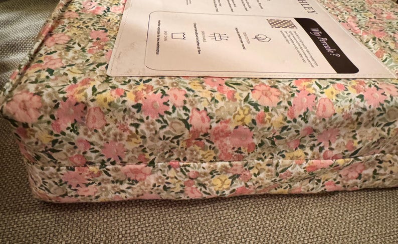 May include: A floral patterned fabric, featuring pink, yellow, and green flowers on a white background. The fabric is folded and appears to be part of a bedding set. A white label is visible on top.