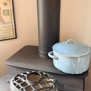 May include: A light blue enamel pot with a lid and handles sits on a black surface. A black metal stove pipe is behind the pot. A decorative metal grate is in the foreground.