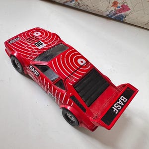 May include: A red toy car with white concentric circle designs on the hood and roof. The car has the number "08" and the word "BASF" printed on it. The car has black wheels and a black spoiler.