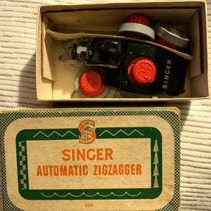 May include: A vintage Singer Automatic Zigzagger sewing machine attachment in its original box. The box is green and white with the words "Singer Automatic Zigzagger" printed on it. The attachment is black with red and silver accents.