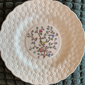 May include: White ceramic saucer with a raised floral design and a central image of a colorful floral tree branch.
