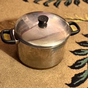 May include: A stainless steel cooking pot with a lid and two black handles. The pot has a round shape and a black knob on the lid. The pot is sitting on a brown surface.