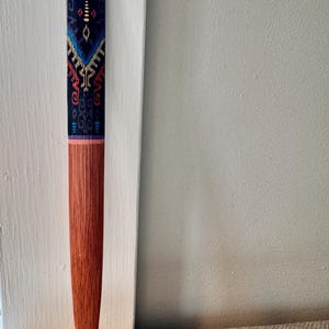 May include: A wooden letter opener with a patterned design. The top portion features a dark blue background with a colorful geometric pattern in blue, red, and yellow. The bottom is a reddish-brown color.