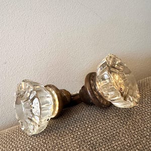 May include: Two antique clear glass door knobs with a faceted design. The knobs have a brass base and are resting on a textured, neutral-colored surface. The glass has a starburst pattern.