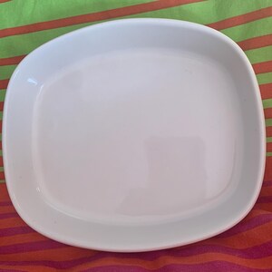 May include: A white ceramic baking dish with a square shape and slightly rounded edges. The dish is empty and has a smooth, glossy finish.