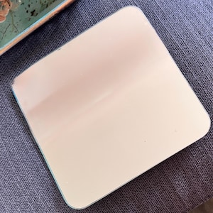 May include: A square, off-white mirror with rounded corners. The mirror has a light blue border and is set against a textured, gray background. The image is a close-up of the mirror.