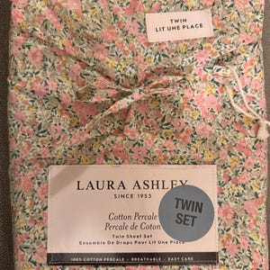 May include: A folded twin sheet set with a floral pattern in pink, yellow, and green. The packaging includes a label that reads "LAURA ASHLEY SINCE 1953" and "TWIN SET". The set is made of 100% cotton percale.