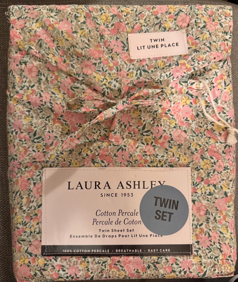 May include: A folded twin sheet set with a floral pattern in pink, yellow, and green. The packaging includes a label that reads "LAURA ASHLEY SINCE 1953" and "TWIN SET". The set is made of 100% cotton percale.