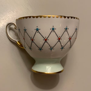 Puede incluir: Taza de t&eacute; blanca con borde y asa dorados, con un patr&oacute;n geom&eacute;trico de l&iacute;neas negras, puntos azules y naranjas. La base es de color verde claro y la taza tiene un borde dorado festoneado.