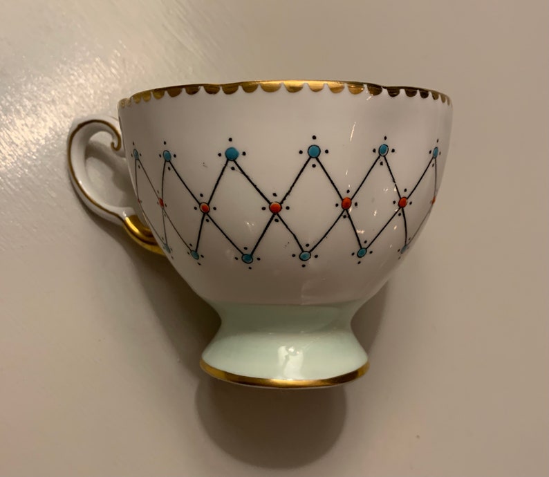 Puede incluir: Taza de t&eacute; blanca con borde y asa dorados, con un patr&oacute;n geom&eacute;trico de l&iacute;neas negras, puntos azules y naranjas. La base es de color verde claro y la taza tiene un borde dorado festoneado.