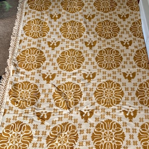 May include: A rectangular textile with a repeating pattern of stylized floral and bird motifs in gold against an off-white background. The textile has a fringed edge. The overall design is symmetrical and ornate, suggesting a decorative or home decor item.