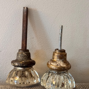 May include: Two vintage glass door knobs with brass-colored bases. One knob has a dark, rusty screw, while the other has a silver screw. The glass is clear and ribbed, and the knobs show signs of age.
