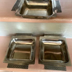 May include: Three silver serving dishes with ornate decorative handles. The dishes are square and have a shiny finish.