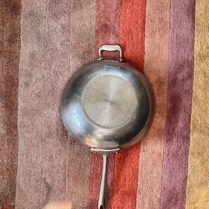 May include: A stainless steel wok-style pan with a long handle and a smaller loop handle. The pan has a rounded bottom and a shiny, reflective surface. The handle is attached with rivets. The pan is sitting on a striped, multi-colored surface.