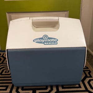 May include: A white and blue Igloo Playmate Maxcold cooler. The cooler has a white lid with a handle and a blue base. The Playmate Maxcold logo is displayed on the front of the cooler.