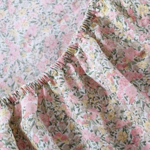 May include: Close-up of a fitted sheet with a floral pattern. The fabric is white with a repeating design of pink, yellow, and green flowers and leaves. The elasticized edge of the sheet is visible.