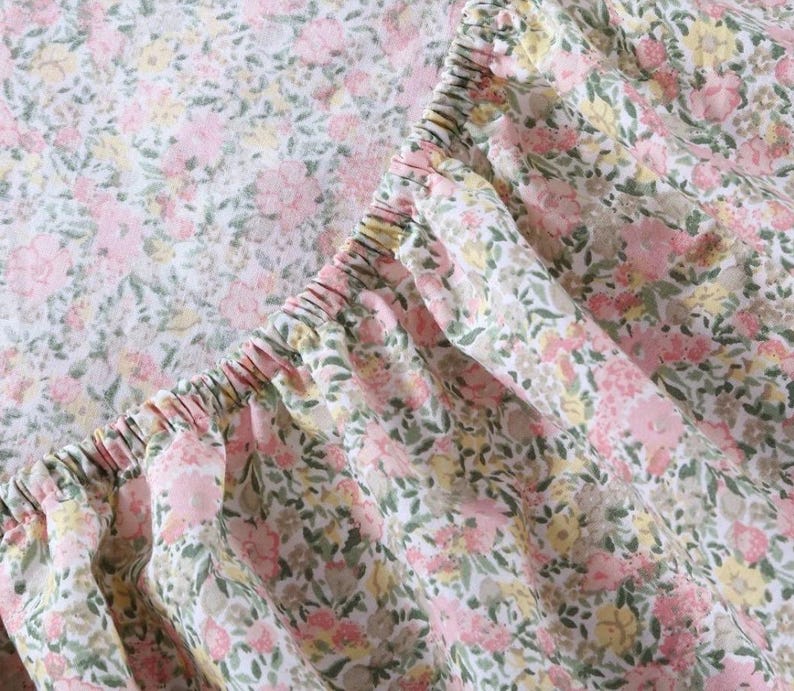 May include: Close-up of a fitted sheet with a floral pattern. The fabric is white with a repeating design of pink, yellow, and green flowers and leaves. The elasticized edge of the sheet is visible.