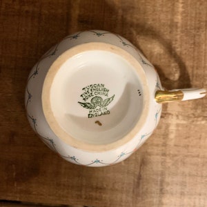 Puede incluir: Taza de porcelana fina inglesa blanca con asa y borde dorados. La taza presenta un delicado estampado floral con detalles azules y rojos. La base de la taza tiene un sello verde que dice "Tuscan Fine English Bone China Made in England."