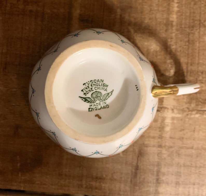 Puede incluir: Taza de porcelana fina inglesa blanca con asa y borde dorados. La taza presenta un delicado estampado floral con detalles azules y rojos. La base de la taza tiene un sello verde que dice "Tuscan Fine English Bone China Made in England."