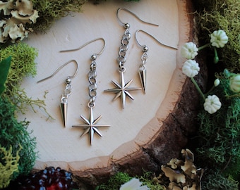 May include: Silver-toned earrings with star and spike designs. The earrings are displayed on a wooden surface surrounded by moss, berries, and white flowers. The earrings have a hook closure.