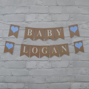 May include: A banner with the words "BABY LOGAN" in wooden letters, strung across a white brick wall. The banner is made of brown paper pennants with light blue heart accents.