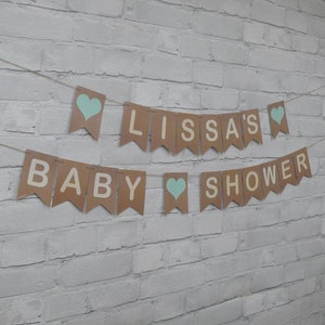May include: A brown paper banner with the words "Baby Lissa's Shower" in white letters and blue hearts. The banner is strung up on a white brick wall.