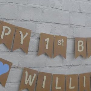 Op de afbeelding: Een bruine papieren banner met blauwe hartjes en de tekst "Happy 1st Birthday William".