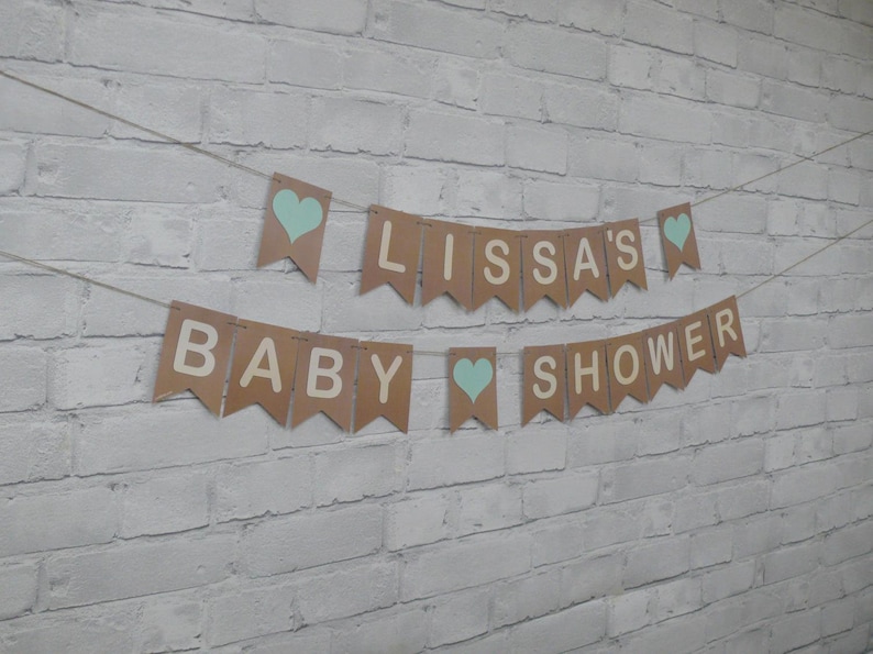 May include: A brown paper banner with the words "Baby Lissa's Shower" in white letters and blue hearts. The banner is strung up on a white brick wall.