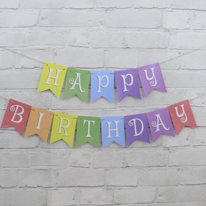May include: A colorful "Happy Birthday" banner with individual pennants in rainbow hues. Each pennant displays a letter in white, strung together with twine. The banner is set against a white brick wall.