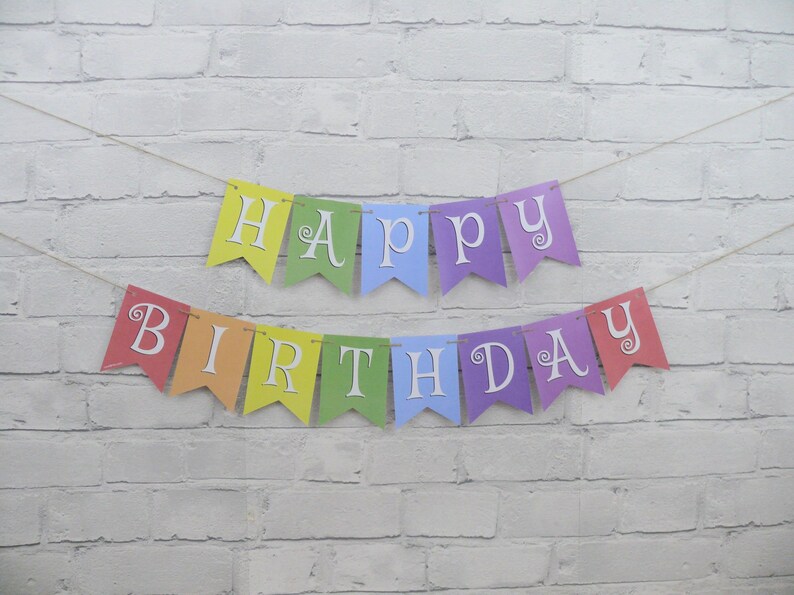 May include: A colorful "Happy Birthday" banner with individual pennants in rainbow hues. Each pennant displays a letter in white, strung together with twine. The banner is set against a white brick wall.