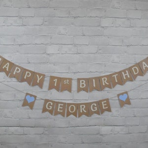 Op de afbeelding: Een bruine papieren banner met blauwe hartjes en de tekst "Happy 1st Birthday George" hangt aan een witte bakstenen muur.