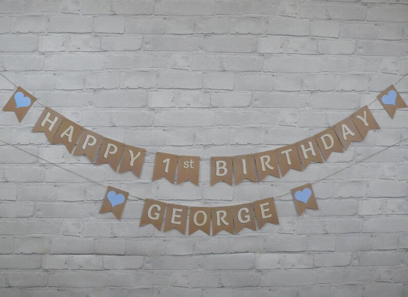 Op de afbeelding: Een bruine papieren banner met blauwe hartjes en de tekst "Happy 1st Birthday George" hangt aan een witte bakstenen muur.