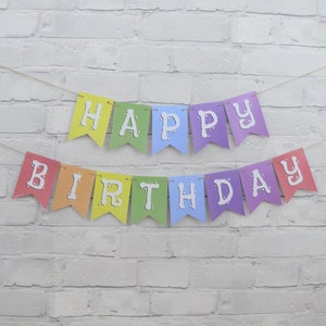 May include: A colorful "Happy Birthday" banner with rainbow-colored pennants. Each pennant displays a letter in white, strung on a thin, natural-colored twine. The banner is set against a white brick wall.