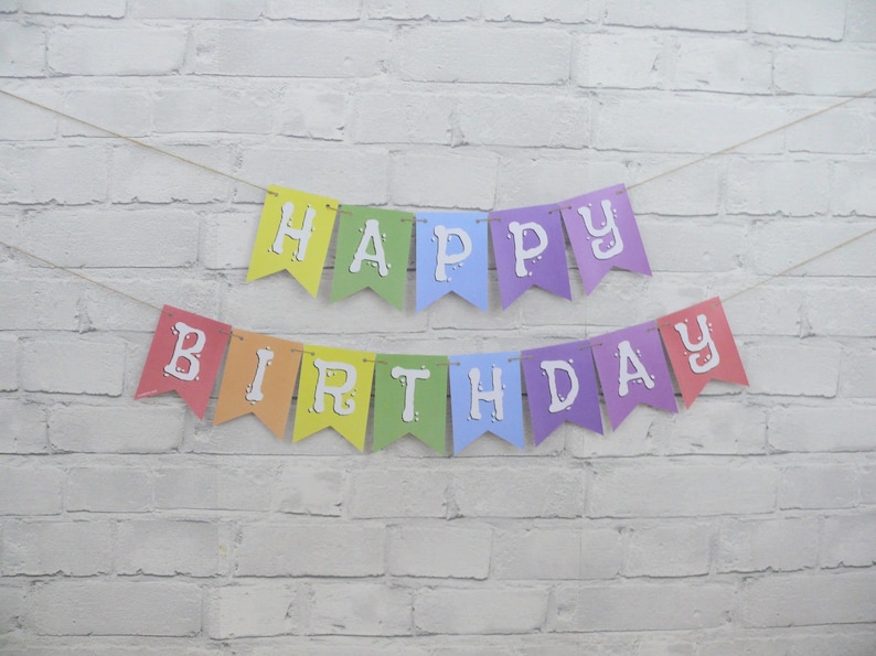 May include: A colorful "Happy Birthday" banner with rainbow-colored pennants. Each pennant displays a letter in white, strung on a thin, natural-colored twine. The banner is set against a white brick wall.