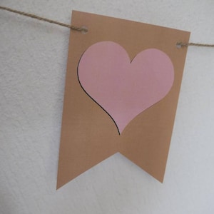 May include: A brown banner with a pink heart cutout, strung on a natural twine. The banner has a rectangular shape with a pointed bottom. The heart is centered on the banner and has a black outline.
