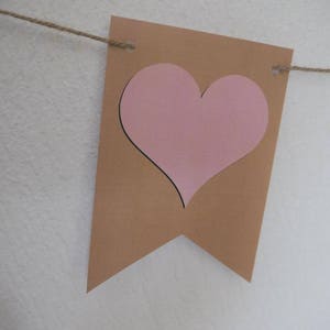 Op de afbeelding: Een bruine papieren banner met een uitgesneden roze hart. De banner is vastgemaakt met touw.