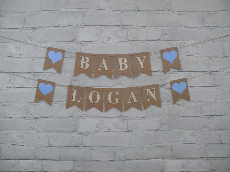 May include: A banner with the words "BABY LOGAN" in wooden letters, strung across a white brick wall. The banner is made of brown paper pennants with light blue heart accents.