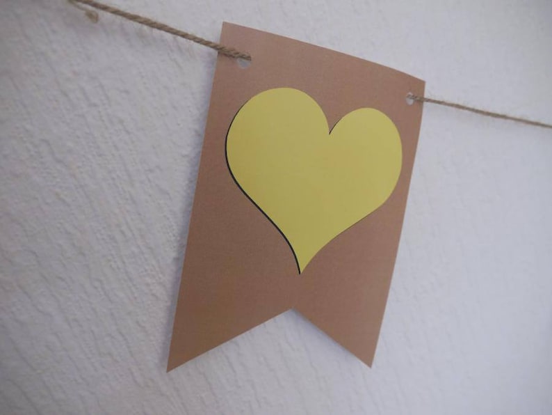 May include: A brown banner with a yellow heart cutout, strung on a natural twine. The banner is made of cardstock and has a simple, minimalist design. The heart is centered on the banner, creating a focal point. Perfect for a party or event.