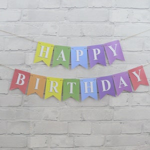May include: A colorful "Happy Birthday" banner with individual pennants in rainbow hues. Each pennant displays a white letter, strung together on a thin twine, against a white brick wall background. The banner is designed for a birthday celebration.