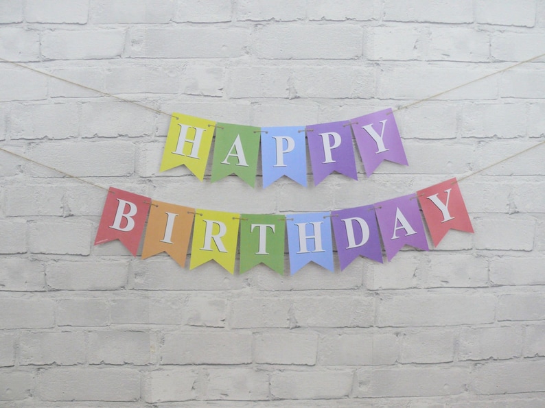 May include: A colorful "Happy Birthday" banner with individual pennants in rainbow hues. Each pennant displays a white letter, strung together on a thin twine, against a white brick wall background. The banner is designed for a birthday celebration.