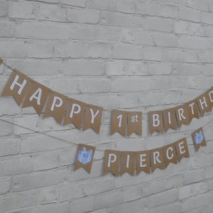Op de afbeelding: Een bruine banner met witte tekst "HAPPY 1st BIRTHDAY PIERCE". De banner is versierd met blauwe cadeau-illustraties en geregen op touw, ideaal voor een eerste verjaardag.
