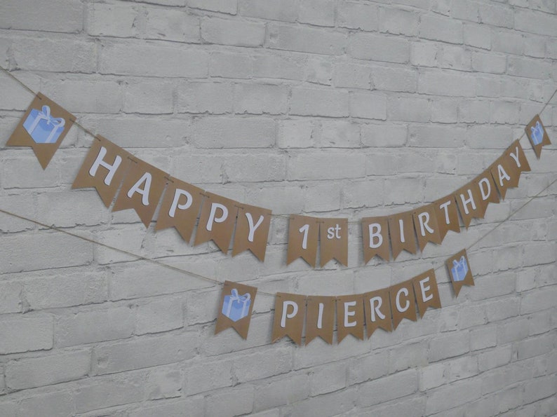 Op de afbeelding: Een bruine banner met witte tekst "HAPPY 1st BIRTHDAY PIERCE". De banner is versierd met blauwe cadeau-illustraties en geregen op touw, ideaal voor een eerste verjaardag.